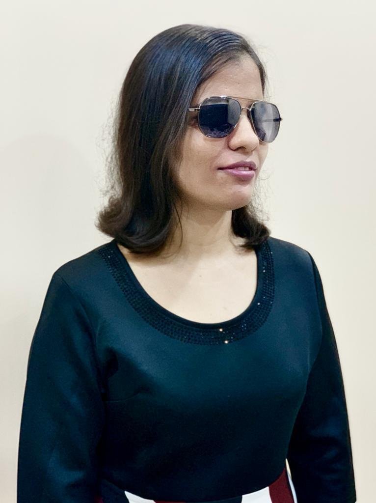 A close-up portrait of Neha wearing dark aviator-style sunglasses and a dark long-sleeved top against a plain light wall.