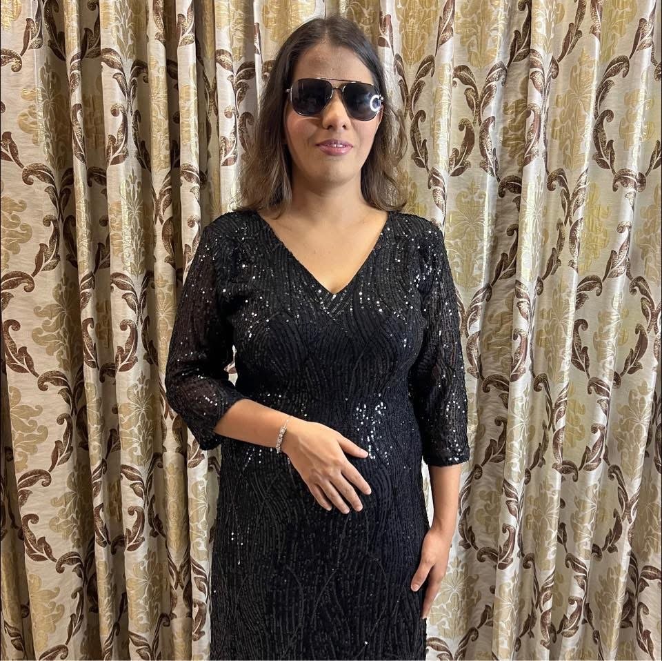 Neha standing in front of gold-patterned curtains, wearing dark sunglasses and a black sequined v-neck dress, posing with one hand placed over her waist.