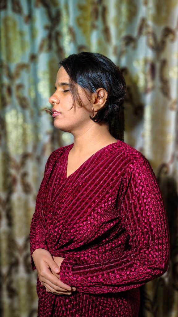 Neha posing in a side profile wearing a long-sleeved, deep burgundy or magenta textured outfit with her hands resting together in front of patterned curtains.