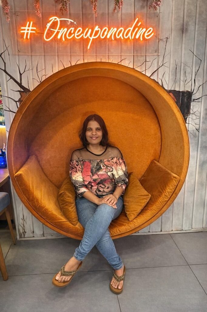 Neha sitting cross-legged inside a large, circular orange pod-style armchair under a bright neon sign that reads '#Onceuponadine'. She is wearing a floral top and blue jeans.