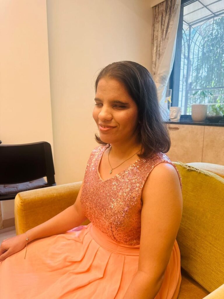 Neha wearing a peach dress, sitting gracefully on a yellow armchair indoors while smiling softly off-camera.